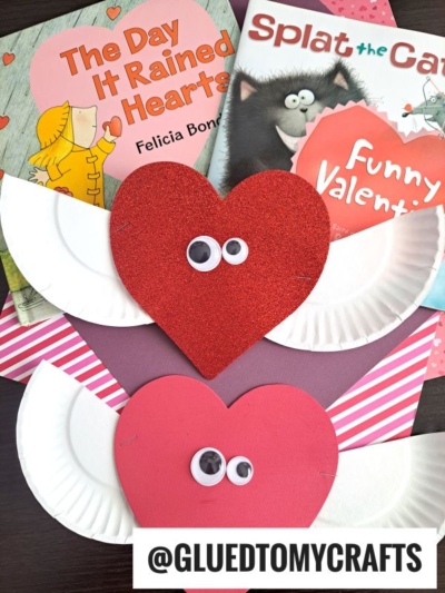 Two Valentines Day-themed children’s books, heart-shaped crafts with googly eyes made from craft foam, and paper plate wings are arranged on patterned paper. One heart is red and glittery, the other is solid pink—fun valentine art project ideas for kids.