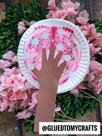 A paper plate decorated like a cherry blossom tree with pink colors and white dots, featuring a brown paper cutout of a hand as the trunk. This PAINT SPLAT CRAFTS project is surrounded by pink flowers. @GLUEDTOMYCRAFTS is written at the bottom.