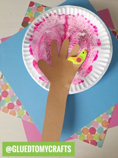 A paper hand cutout with a small bird on the finger is glued to a pink-painted paper plate, placed on colorful scrapbook paper—perfect for fans of PAINT SPLAT CRAFTS. The image has a green @GLUEDTOMYCRAFTS label at the bottom.