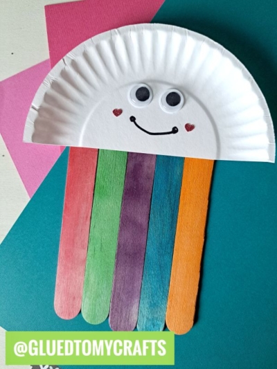 A rainbow and cloud craft made from a paper plate half with googly eyes, a smiling face, and red paper hearts, with colorful craft sticks as the rainbow rays, on a teal background. @gluedtomycrafts is labeled at the bottom.