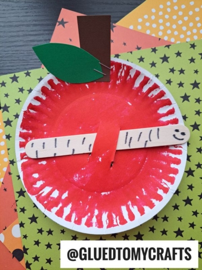 A paper plate painted red with a craft stick glued across it, resembling an apple. Green paper leaves and a brown paper stem are attached at the top. Great for INSECT CRAFT IDEAS, the craft stick has a face and black lines drawn on it.