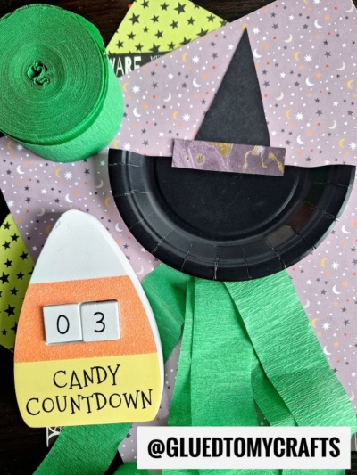 A witch paper plate craft with green streamers, a candy corn countdown showing “03,” and rolls of crepe paper—perfect for your next crepe paper craft ideas—are displayed on star-patterned paper. Text at the bottom reads @GLUEDTOMYCRAFTS.