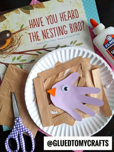 A children’s craft setup features a paper plate with a hand-shaped purple cutout and brown paper strips resembling a nest, purple scissors, glue, and the book Have You Heard the Nesting Bird? in the background.