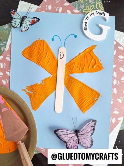 A kids’ butterfly craft features orange paint wings and a craft stick with a smiley face as the body, placed on blue paper with faux butterflies, patterned paper in the background, and a pie server stamped “@GLUEDTOMYCRAFTS” handle.
