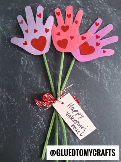 Three handprint-shaped paper flowers with red heart decorations are attached to green stems and tied with a red polka-dot ribbon. A tag reads Happy Valentines Day! All are set on a dark surface.