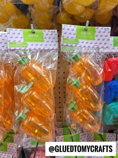 Plastic carrot-shaped Easter eggs with green tops are displayed in hanging packages on a store shelf. The packaging has a pastel bunny design and is labeled “Easter Carrot Shaped Eggs.” Price tags show $2.99 each.