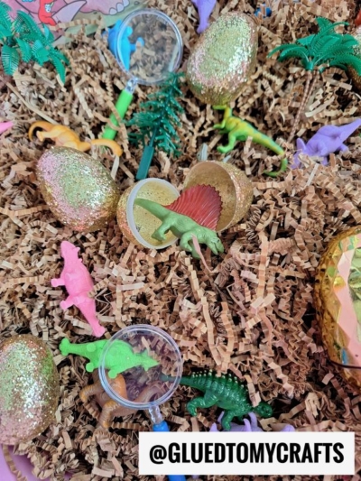 Toy dinosaurs, glittery gold eggs, magnifying glasses, and small plastic trees are scattered on brown crinkled paper shreds. The Instagram handle @GLUEDTOMYCRAFTS is displayed at the bottom.