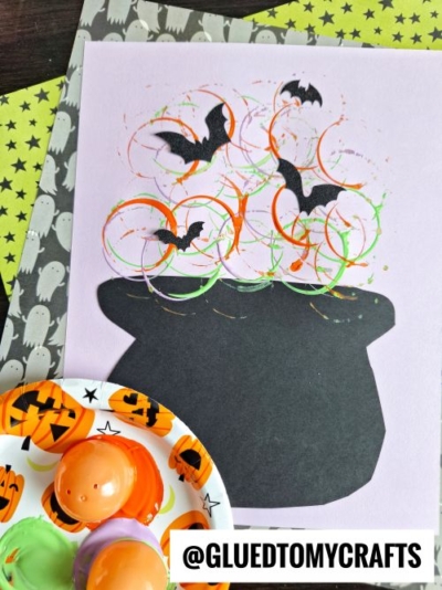 A Halloween craft features a black cauldron on purple paper, with painted orange, green, and black swirls made using Plastic Egg Stamp Crafts and bat cutouts above it. Pumpkin and ghost-themed items are nearby. Text reads @GLUEDTOMYCRAFTS at the bottom.
