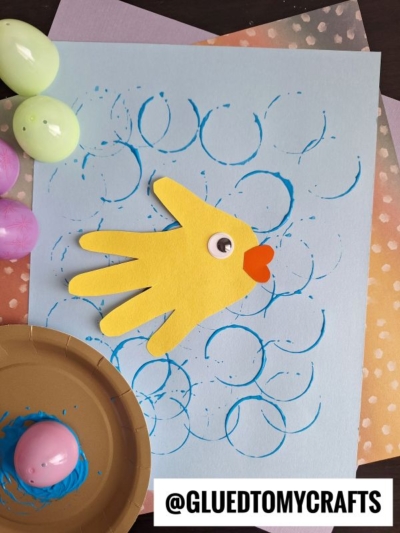 A yellow paper fish made from a handprint cutout with a googly eye and orange mouth is glued on blue paper, surrounded by blue circle prints created using Plastic Egg Stamp Crafts. Pastel eggs and craft supplies are nearby.