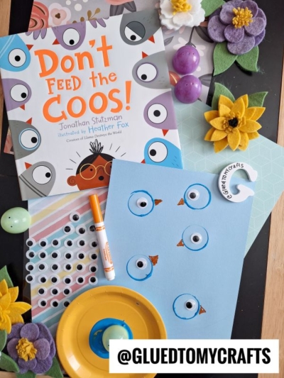 A children’s book titled Don’t Feed the Coos! sits on a table with craft supplies for Plastic Egg Stamp Crafts, including construction paper, a marker, a sunflower, purple eggs, a yellow pom-pom flower, and blue paper with drawn bird faces.