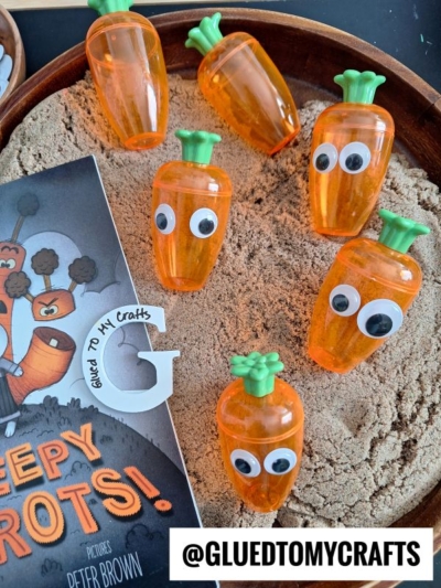 Plastic carrot-shaped containers with googly eyes are arranged on sand next to a childrens book titled Creepy Carrots! and a small wooden G with Glued to My Crafts written on it. @GLUETOMYCRAFTS is written below.