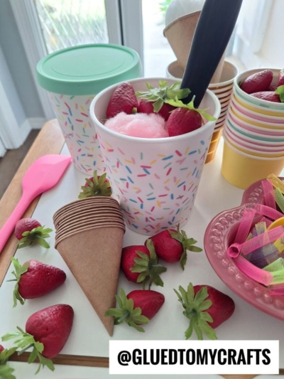 Colorful paper cups with sprinkles design are filled with strawberries and pink cotton candy. Brown paper cones, fresh strawberries, and a pink spatula are also on the table. @GLUEDTOMYCRAFTS is written at the bottom.