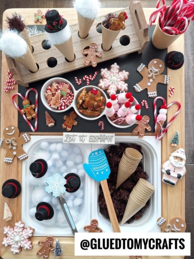 Festive holiday craft setup with gingerbread, snowman, Santa, candy canes, and assorted decorations on a wooden tray—plus playful touches like cones for pom pom ice cream play, a mitten spatula, Let it Snow sign, and neatly arranged treats.