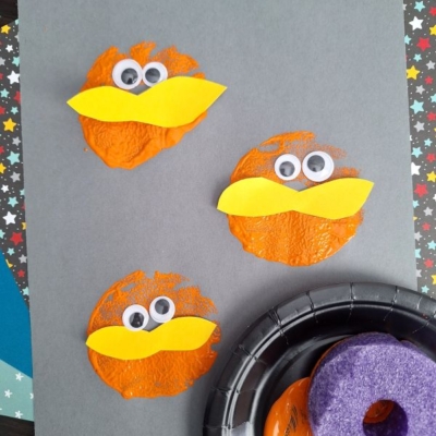 Three orange textured circles on gray paper, each with two googly eyes and a yellow beak made of paper, resembling birds or cartoon characters. A black tray with orange paint and a purple sponge is nearby.