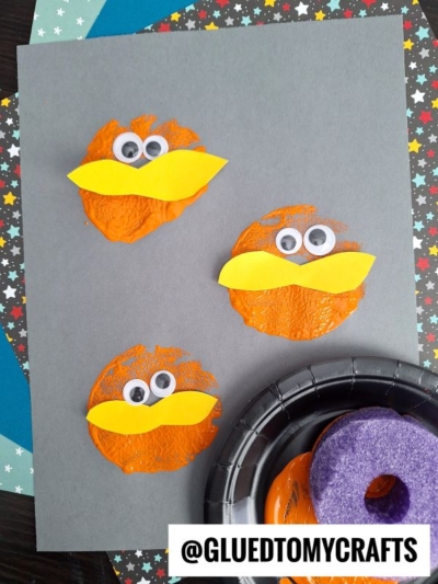 Three orange textured circles on gray paper, each with two googly eyes and a yellow beak made of paper, resembling birds or cartoon characters. A black tray with orange paint and a purple sponge is nearby.