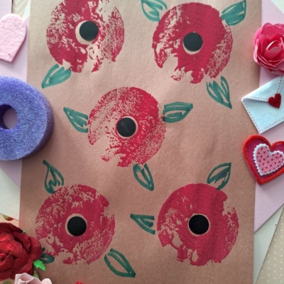 A handmade artwork of five red flowers with green leaves on brown paper, surrounded by felt hearts, a purple ring, and red and pink craft decorations. @GLUEDTOMYCRAFTS is written at the bottom.
