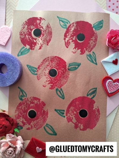 A handmade artwork of five red flowers with green leaves on brown paper, surrounded by felt hearts, a purple ring, and red and pink craft decorations. @GLUEDTOMYCRAFTS is written at the bottom.