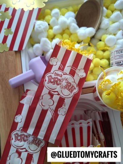 Red and white striped popcorn bags, yellow shredded paper, cotton balls, and craft items arranged on a table. A tag at the bottom reads @GLUEDTOMYCRAFTS. The scene has a playful, crafty feel.