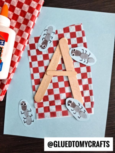 A fun insect craft idea featuring a letter A made from wooden sticks on red checkered paper, surrounded by cartoon ant cutouts, a glue bottle, and a red cloth. @GLUEDTOMYCRAFTS is labeled at the bottom.