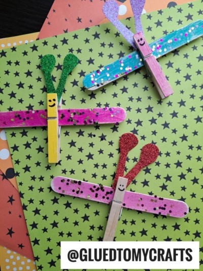 Three colorful dragonflies made from clothespins and popsicle sticks are decorated with glitter and polka dots, placed on green and orange star-patterned paper—perfect for those seeking fun INSECT CRAFT IDEAS. The text @GLUEDTOMYCRAFTS appears at the bottom.
