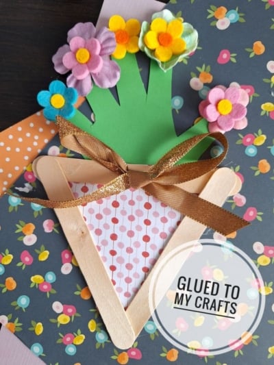 A handmade craft shaped like a bouquet, made from craft sticks, colored paper, felt flowers, and a gold ribbon, displayed on floral-patterned paper with the text Glued To My Crafts.