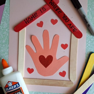 A handmade craft features a house frame made of craft sticks, a handprint cutout with heart shapes, and the phrase “Home is Where You Are.” Elmer’s glue and a marker are nearby. The Instagram handle @GLUEDTOMYCRAFTS is at the bottom.