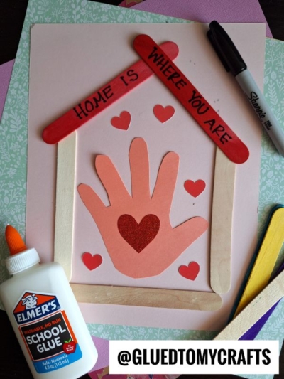 A handmade craft features a house frame made of craft sticks, a handprint cutout with heart shapes, and the phrase “Home is Where You Are.” Elmer’s glue and a marker are nearby. The Instagram handle @GLUEDTOMYCRAFTS is at the bottom.