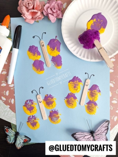 A blue paper displays butterfly crafts made from paint and wooden sticks, surrounded by glue, a sponge, artificial flowers, and butterflies—perfect for INSECT CRAFT IDEAS. The butterfly wings show yellow and purple paint prints. @GLUEDTOMYCRAFTS is written at the bottom.