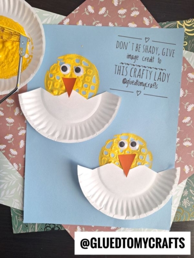 Two potato masher stamped chicks with yellow painted faces, googly eyes, and orange beaks sit on a blue background. Each chick has folded white paper plate wings, with craft supplies and patterned paper nearby.