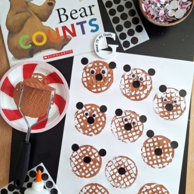 A children’s craft setup features a Bear Counts book, glue, googly eyes, black dot stickers, and a potato masher stamped craft with brown bear faces, plus a finished bear face and supplies displayed on the table.