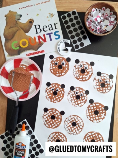 A children’s craft setup features a Bear Counts book, glue, googly eyes, black dot stickers, and a potato masher stamped craft with brown bear faces, plus a finished bear face and supplies displayed on the table.