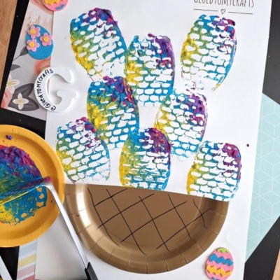 Colorful fish scale prints made with paint and a potato masher cover white paper. Craft supplies, including a gold plate, paint, and foam Easter eggs, are scattered around. @GLUEDTOMYCRAFTS is written at the bottom.