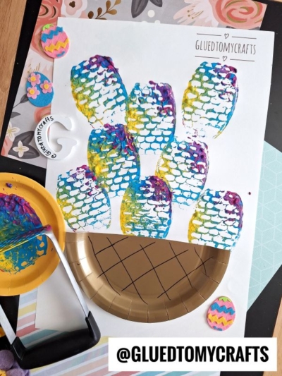 Colorful fish scale prints made with paint and a potato masher cover white paper. Craft supplies, including a gold plate, paint, and foam Easter eggs, are scattered around. @GLUEDTOMYCRAFTS is written at the bottom.