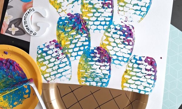 Colorful fish scale prints made with paint and a potato masher cover white paper. Craft supplies, including a gold plate, paint, and foam Easter eggs, are scattered around. @GLUEDTOMYCRAFTS is written at the bottom.