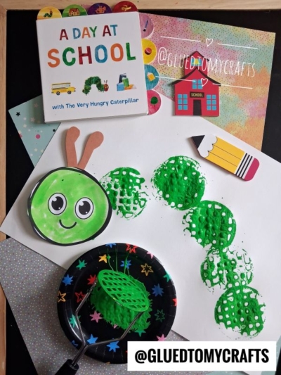 Children’s craft setup inspired by “A Day at School” book: green potato masher stamped caterpillar prints on paper, cute caterpillar face cutout, paint, craft supplies, and school-themed stickers on a colorful background.