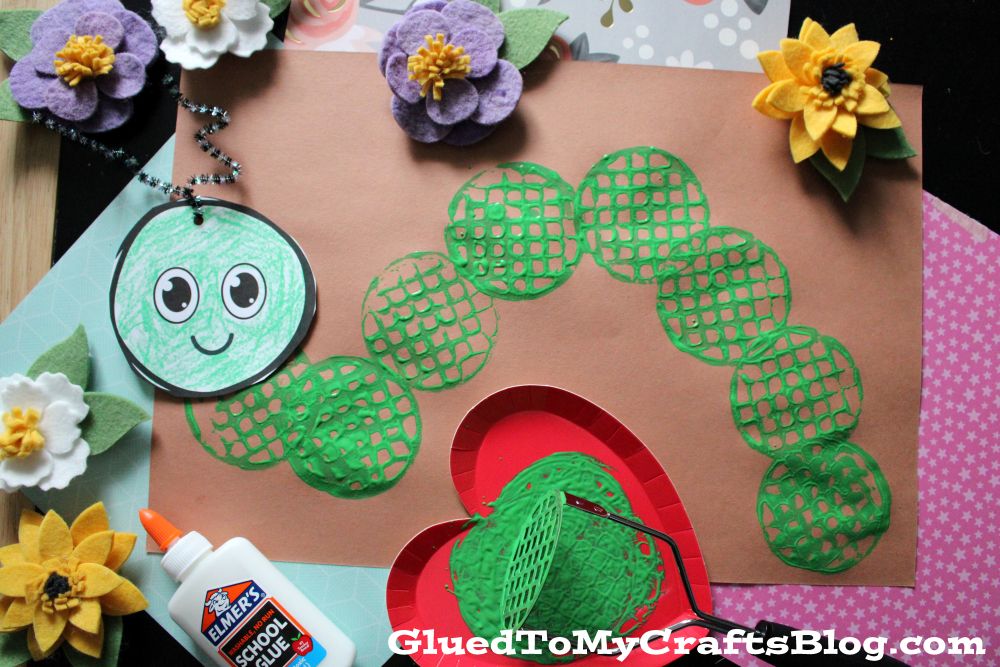 A childrens craft project featuring a green caterpillar made from painted circles on brown paper, surrounded by felt flowers, a bottle of glue, a heart-shaped tray with green paint, and a smiling caterpillar keychain.