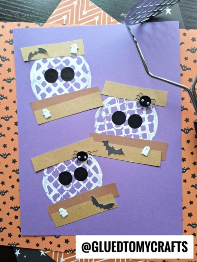 Three mummy faces made from paper, with black eyes, brown paper strips, and Halloween decorations (bats, ghosts, spiders) on a purple background. A potato masher stamped s’mores treat and orange Halloween-themed papers are nearby.