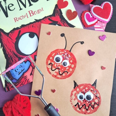 A children’s book titled “Love Monster” is beside a brown craft paper with two red monster faces made from prints, googly eyes, and hearts. Yarn hearts and heart stickers are scattered around. A potato masher is also visible.