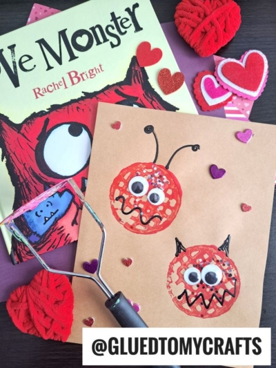 A children’s book titled “Love Monster” is beside a brown craft paper with two red monster faces made from prints, googly eyes, and hearts. Yarn hearts and heart stickers are scattered around. A potato masher is also visible.