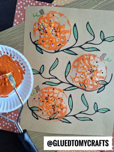 A brown sheet with four pumpkin shapes created using a potato masher stamped craft, painted orange and decorated with glitter and sequins on vine drawings. A paint roller and plate with orange paint are nearby. @GLUEDTOMYCRAFTS is written at the bottom.