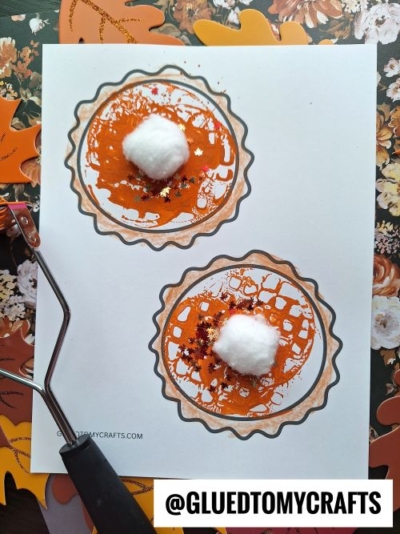 A craft project showing two paper pies decorated with orange paint, glitter, and a cotton ball on top to resemble whipped cream. A paint roller and autumn-themed background are visible in this fun potato masher stamped craft.