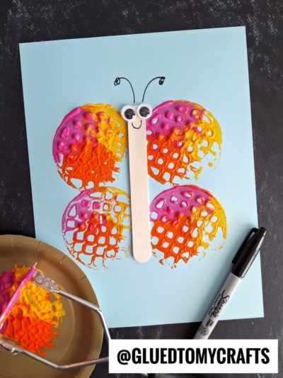 A potato masher stamped butterfly craft featuring four colorful painted waffle-shaped wings and a popsicle stick body with googly eyes and antennae on blue paper. Nearby are a black marker and the painted potato masher used for stamping.