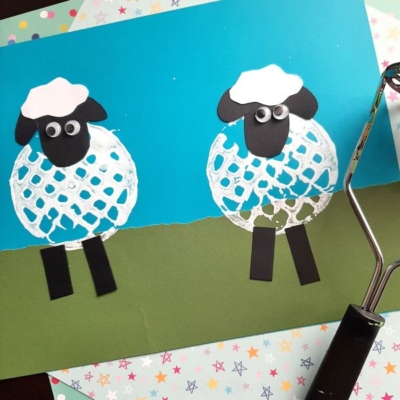 Two sheep crafts made from white paint and paper cutouts are displayed on a blue and green background. A rolling tool is on the right, and colorful star-patterned paper is underneath. @GLUEDTOMYCRAFTS is at the bottom left.