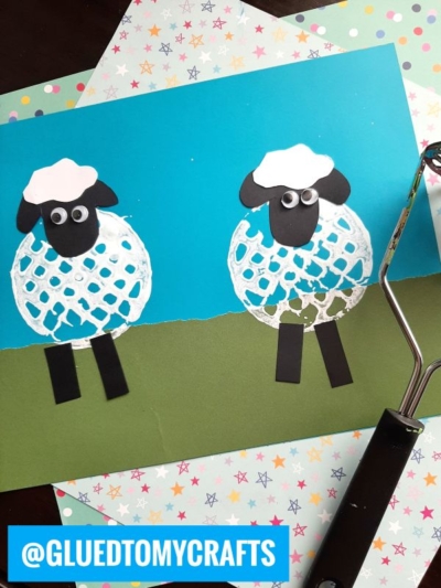 Two sheep crafts made from white paint and paper cutouts are displayed on a blue and green background. A rolling tool is on the right, and colorful star-patterned paper is underneath. @GLUEDTOMYCRAFTS is at the bottom left.