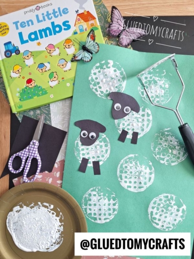 Children’s potato masher stamped sheep craft using white paint circles and black paper sheep on green paper, surrounded by the book Ten Little Lambs, crafting supplies, scissors, paper butterflies, and @GLUEDTOMYCRAFTS label.