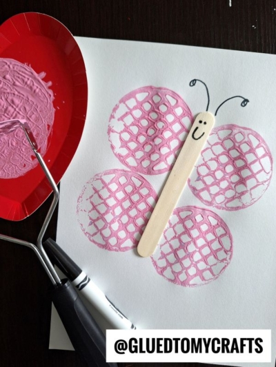 A potato masher stamped butterfly craft made with pink waffle prints for wings and a popsicle stick body, featuring a smiling face and antennae drawn in black marker. A paint roller and plate with pink paint are nearby.