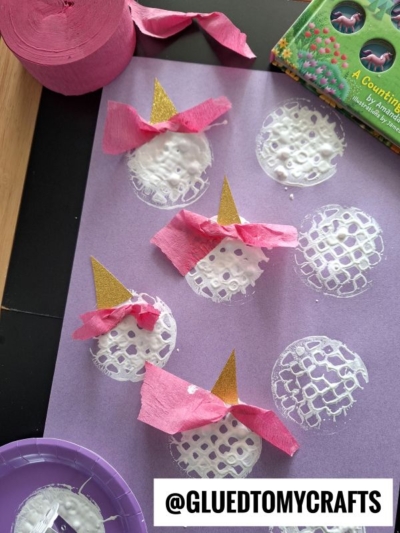 A purple paper displays five unicorn faces made from white textured circles, gold glittery horns, and pink crepe paper bows. Craft supplies and a @GluedToMyCrafts label are also visible.