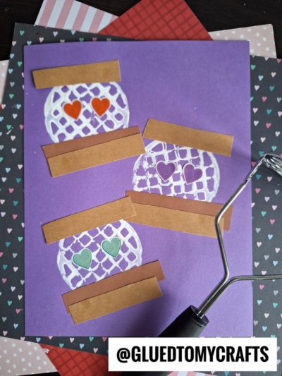 A purple card decorated with three white mason jars, each topped with brown paper strips and containing colored heart shapes, evokes a potato masher stamped s’mores vibe. A black craft tool and patterned paper with hearts surround the card.