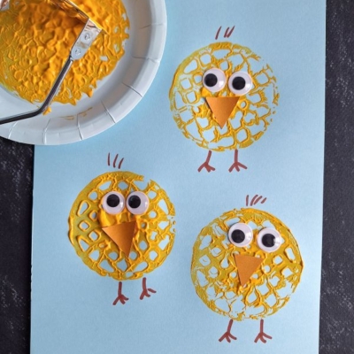 Three yellow chick crafts on blue paper, made with painted bubble wrap circles and one adorable potato masher stamped chick, googly eyes, orange paper beaks, and drawn legs; a paint-covered bubble wrap roller and plate are shown beside them.
