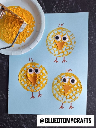 Three yellow chick crafts on blue paper, made with painted bubble wrap circles and one adorable potato masher stamped chick, googly eyes, orange paper beaks, and drawn legs; a paint-covered bubble wrap roller and plate are shown beside them.
